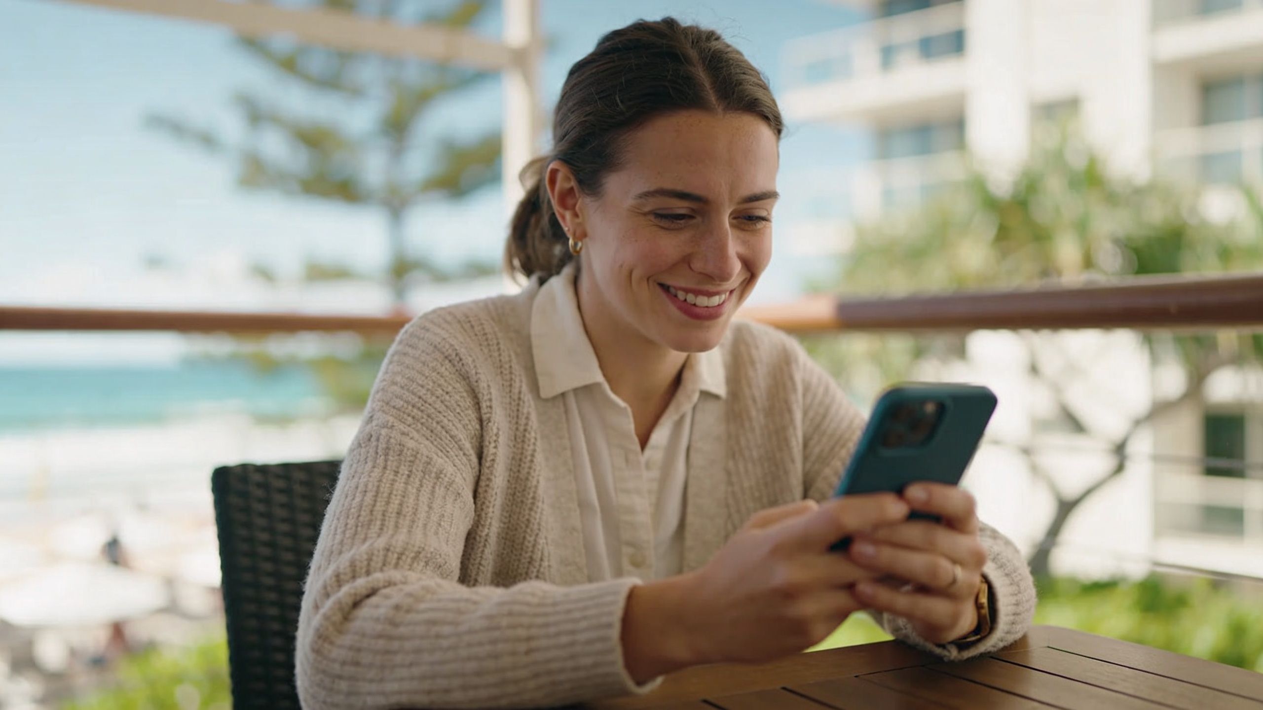 A young woman smiling while looking at her smartphone during a relaxing outdoor break near the ocean.