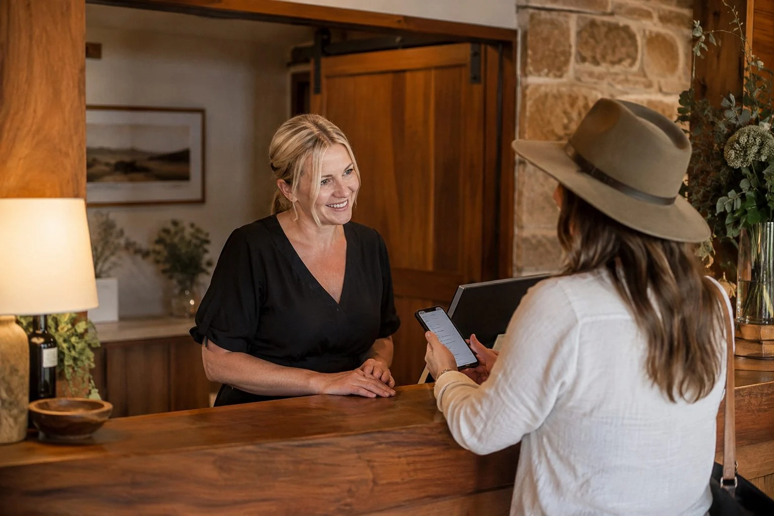 Boutique accommodation owner at her reception desk talking with a guest who is holding up a phone with a question