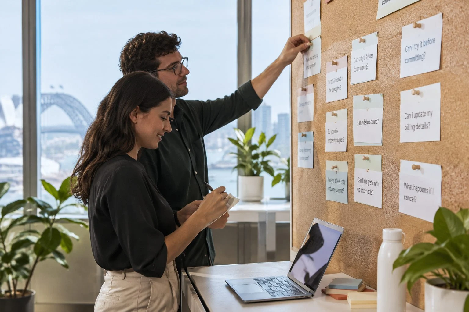 Two colleagues pinning grouped customer questions onto a cork board and writing short answers in a Sydney office